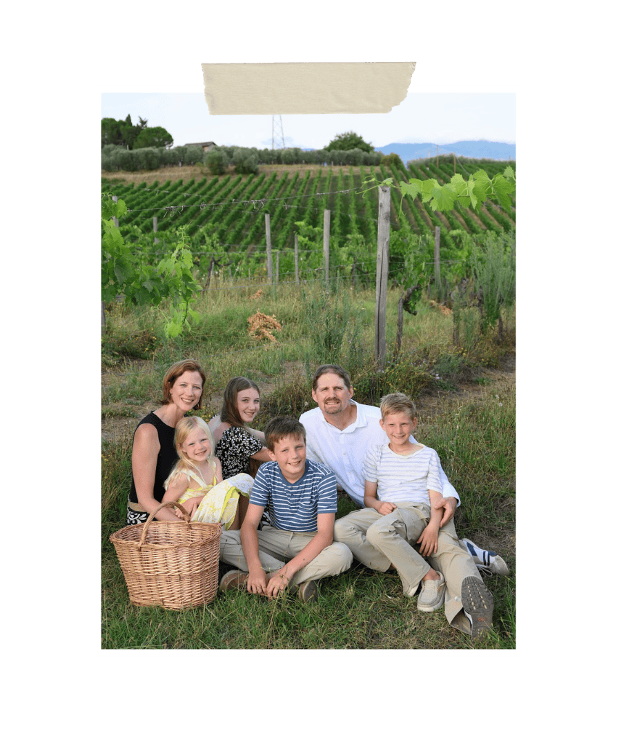 Family of six sitting together in a vineyard with rolling hills in the background, smiling and enjoying a peaceful afternoon outdoors with a picnic basket nearby.