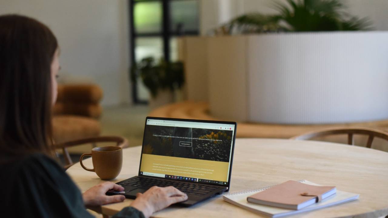A woman sitting at a table using a laptop to access an online psychology course, with a notebook and coffee nearby