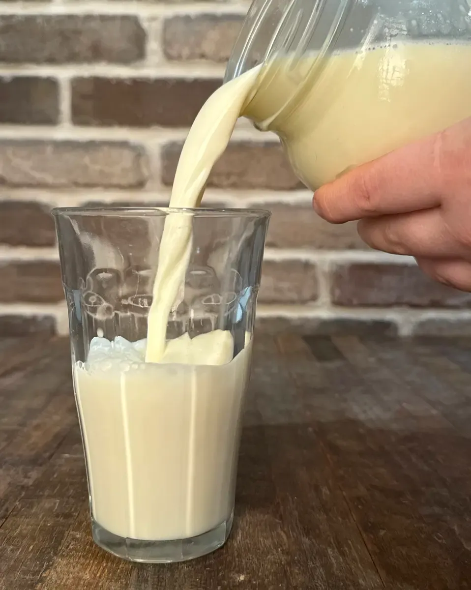 Fresh raw milk being poured from a glass jar into a drinking glass, illustrating milk production and nutrition from cow feed