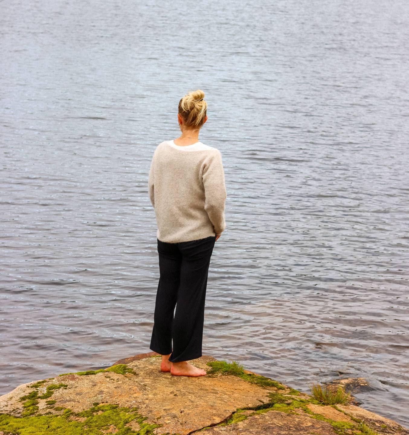 Kvinne i strikkegenser står rolig på en klippe og ser ut over vannet