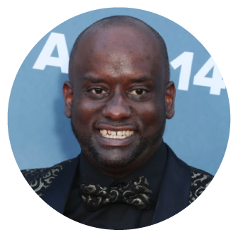 A circle photo headshot of Keith. A Black man with dark skin tone and a shaved head, he is looking at the camera and smiling. He is wearing a gold and black bowtie and tuxedo jacket and is standing in front of a light blue backdrop. 