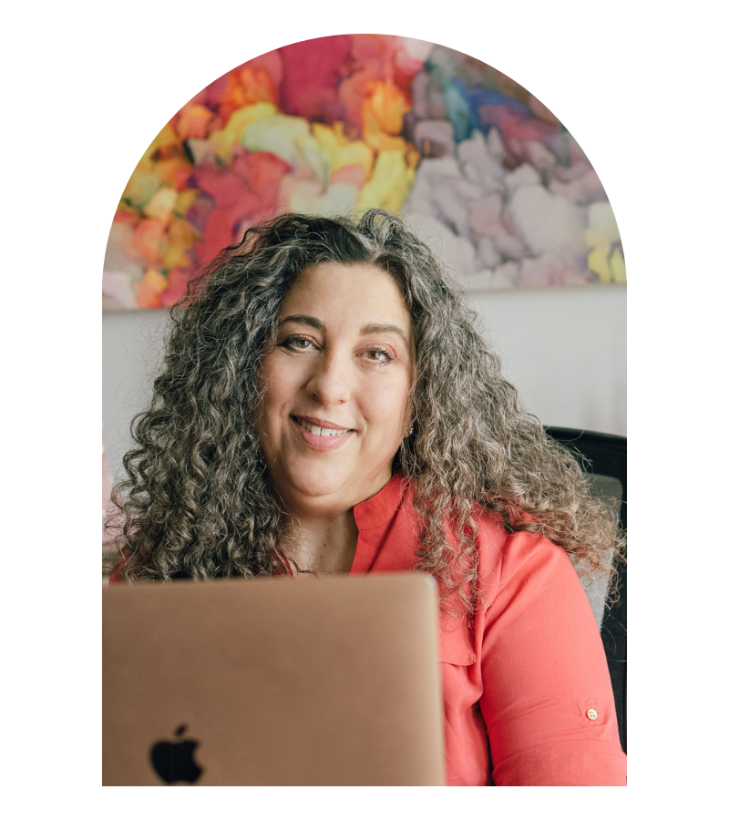 woman with long cirly brown hair at a computer smiling