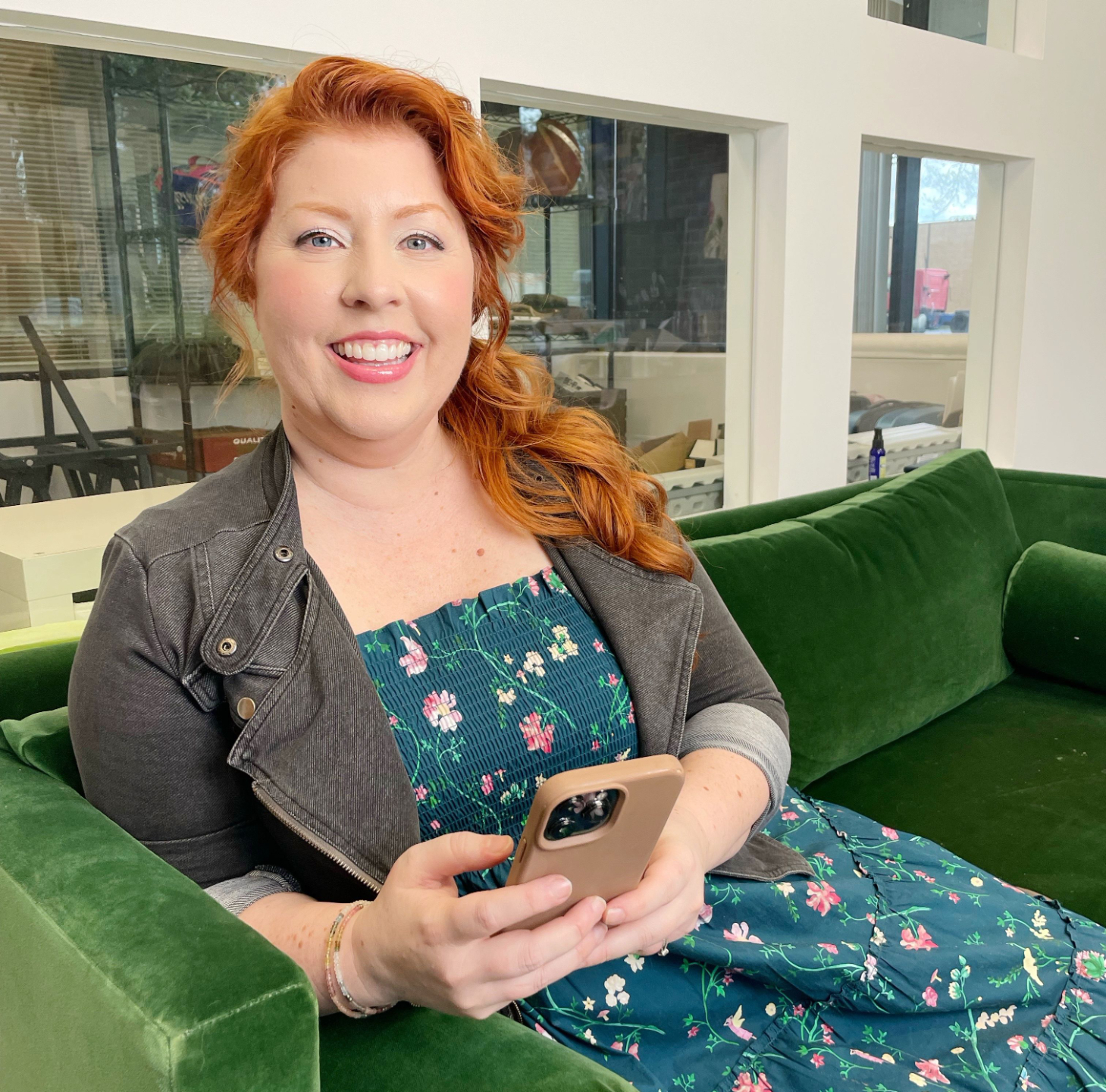 white redheaded woman in green dress with gray denim jacket.  shes's smiling and holding a phone that she's typing on