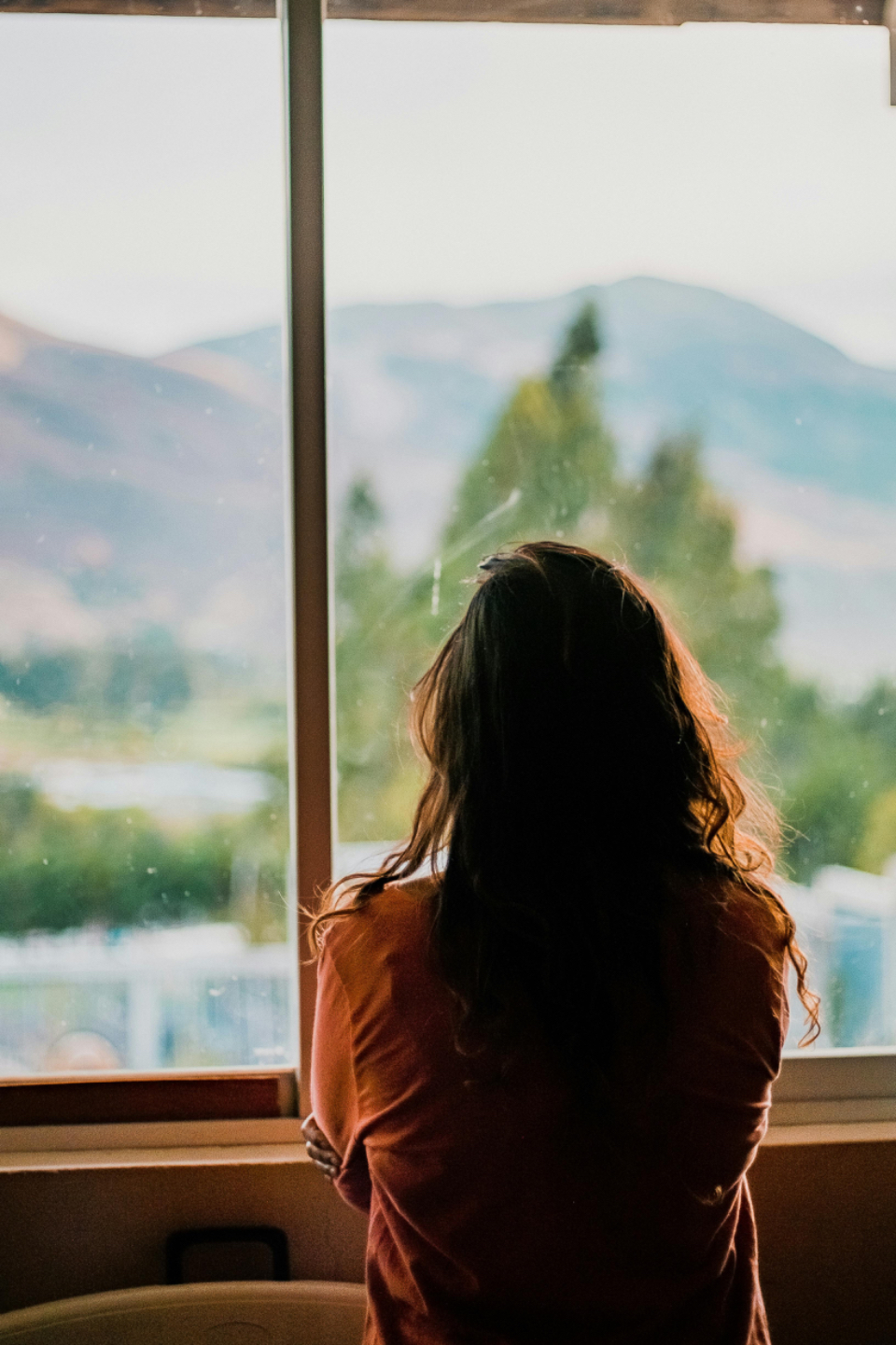 A thoughtful mom looking out a window, reflecting on the emotional challenges of parenting a teen or young adult and seeking support through life coaching for parents