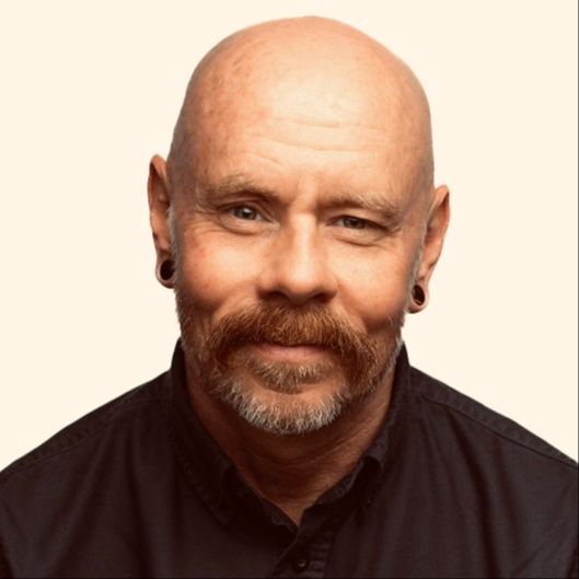 Headshot of smiling, welcoming 50s bald white gay cisgender white man with ginger-grey beard, pierced ears, and black shirt