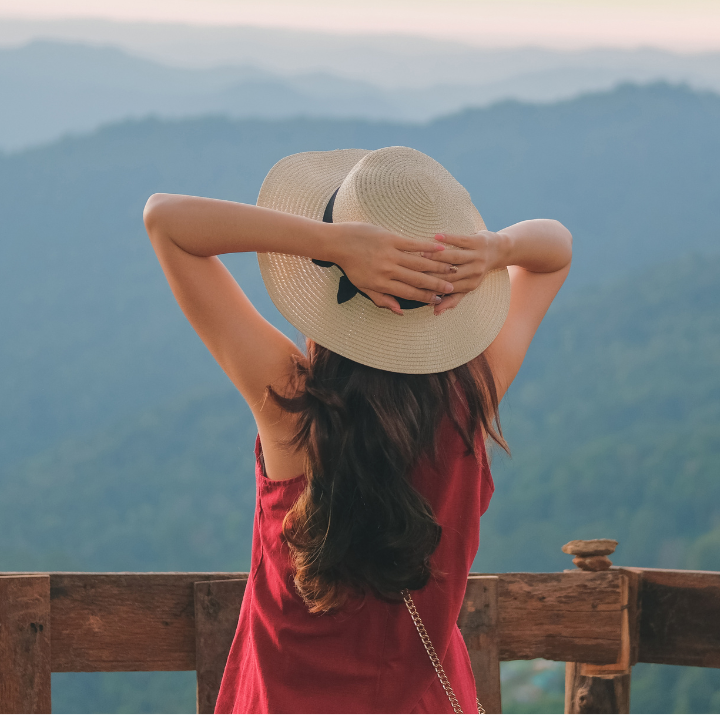 Woman standing on a wooden overlook facing scenic blue mountains, wearing a straw hat and red dress, with hands resting on her head — expressing calm reflection, freedom, and peace from practicing mindfulness and purpose alignment.
