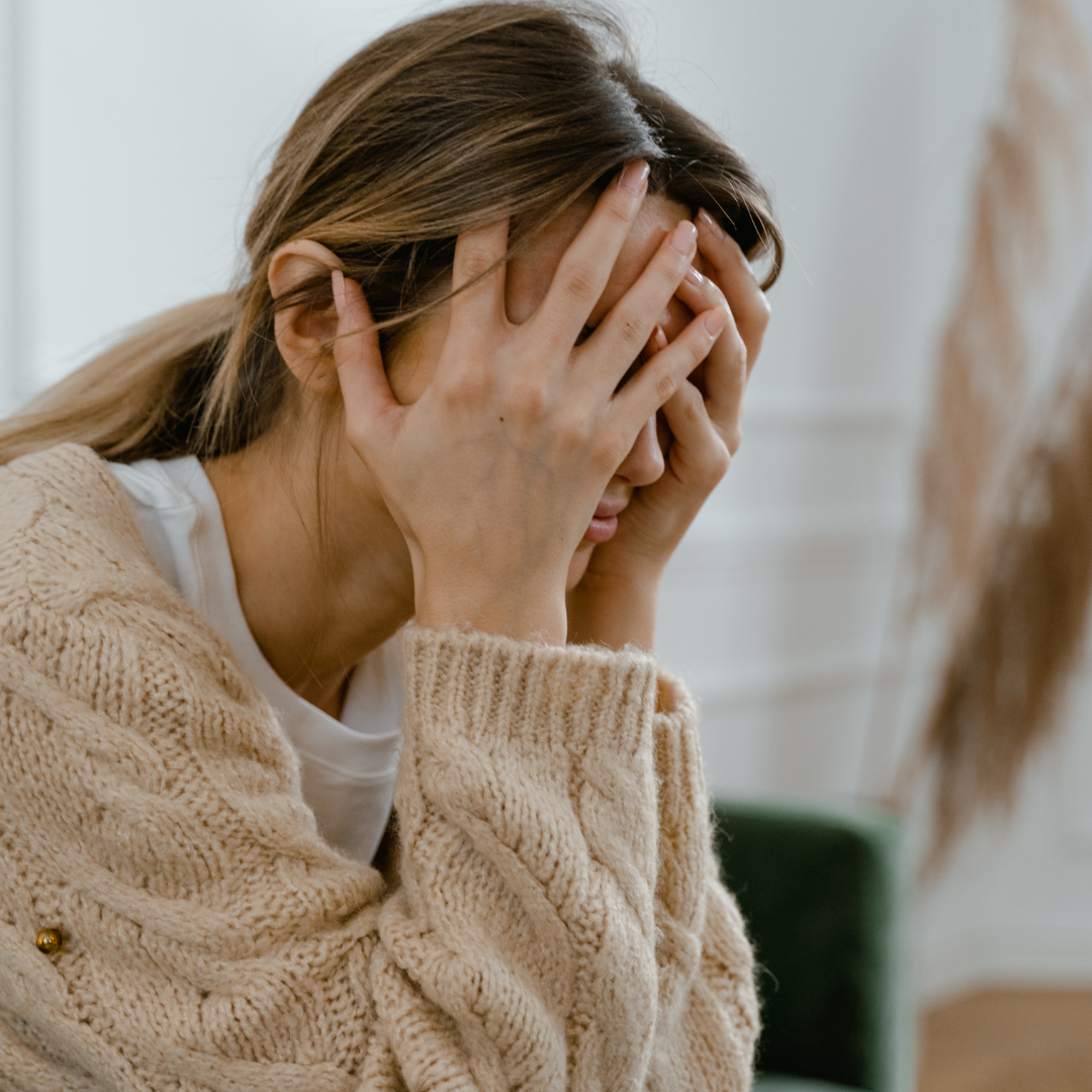 Woman sitting indoors with her hands covering her face, wearing a beige knit sweater — representing feelings of overwhelm, stress, or emotional exhaustion before starting personal growth or self-care work.