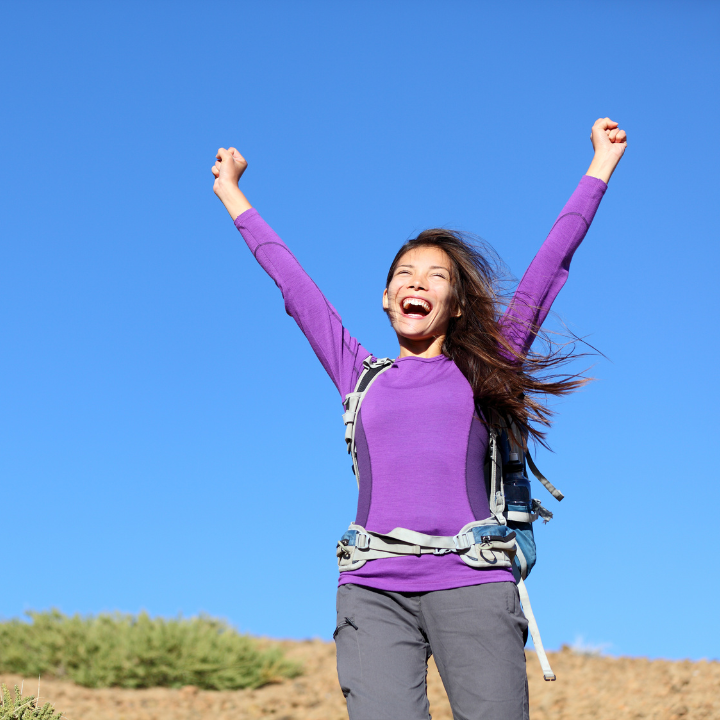 Smiling woman outdoors wearing a purple long-sleeve shirt and hiking gear, raising her arms joyfully toward the blue sky — representing empowerment, confidence, and freedom after completing a mindset transformation.
