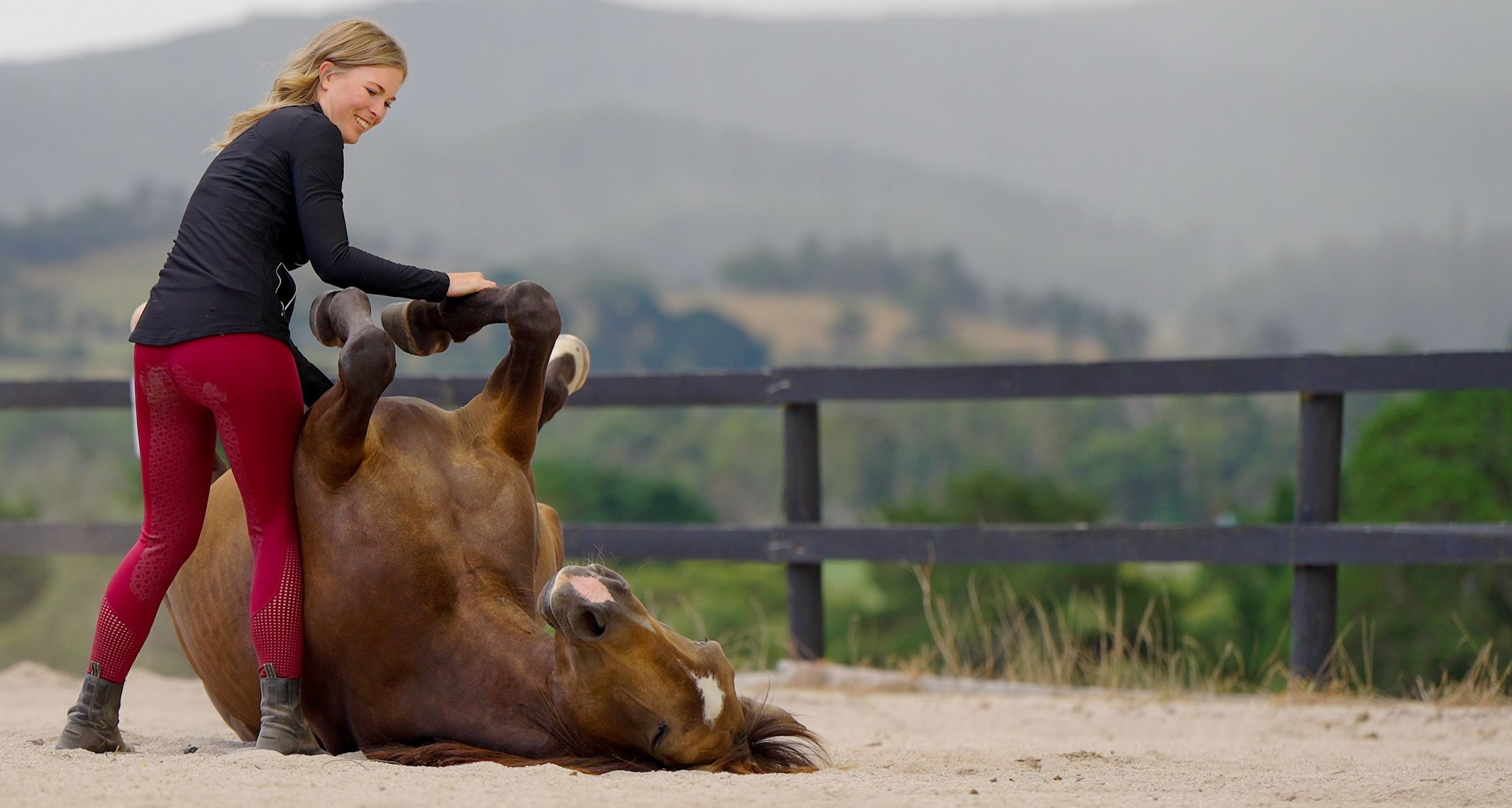 Nadine smiling as a chestnut horse lies on its back beside her in the arena, legs in the air