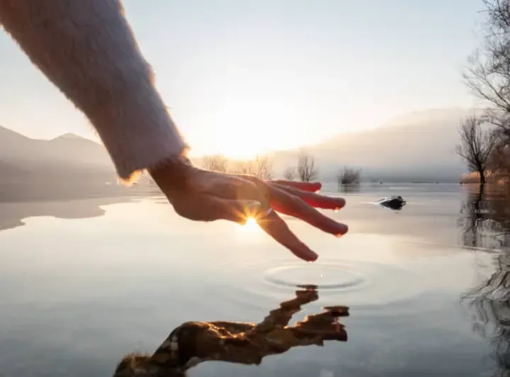 Hand gently touching water at sunrise, symbolizing the self-discovery and reflection in Vivian’s Human Design blueprint.