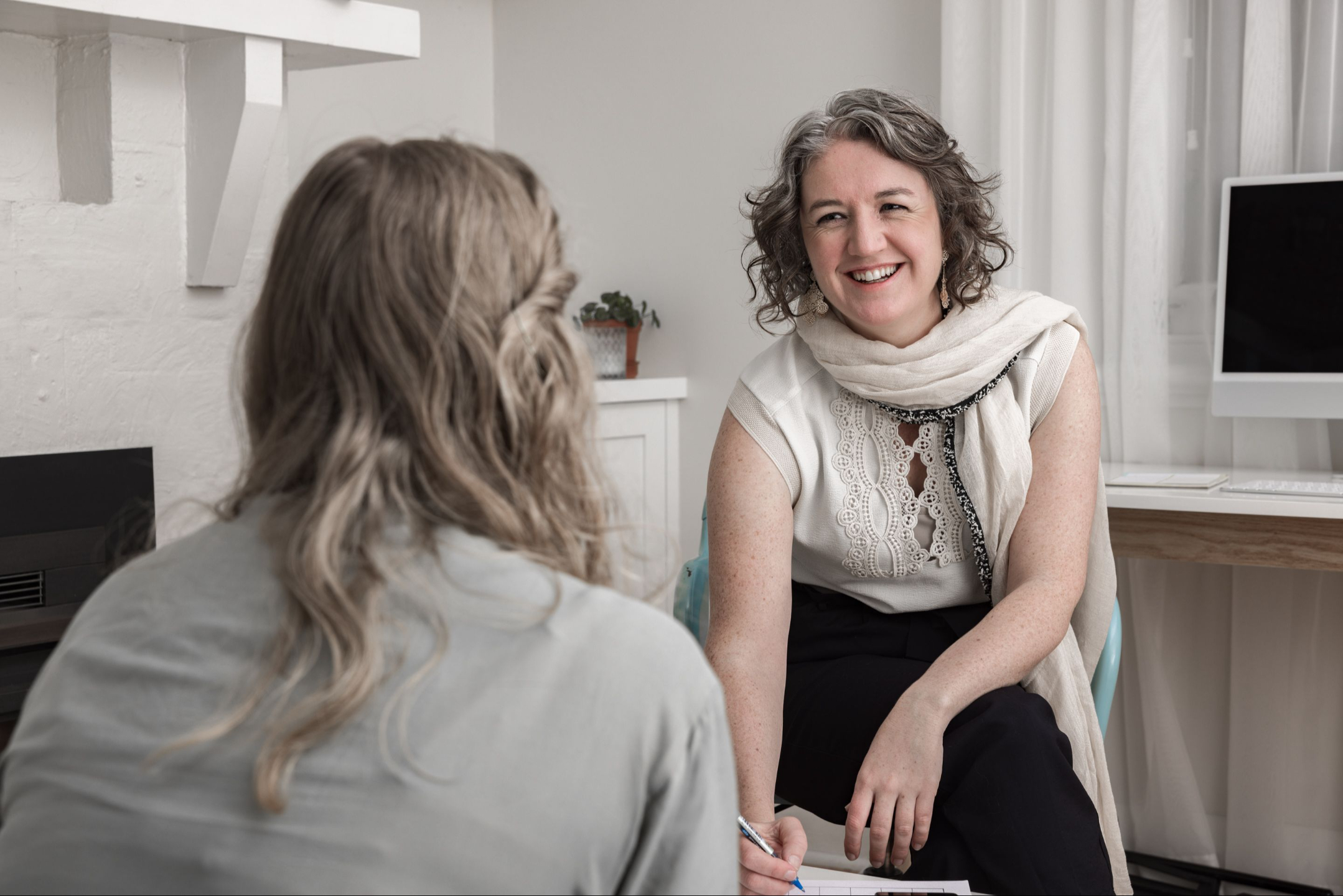 Founder Kate smiling while talking with a client in a light-filled office, holding a pen and leaning forward in conversation.