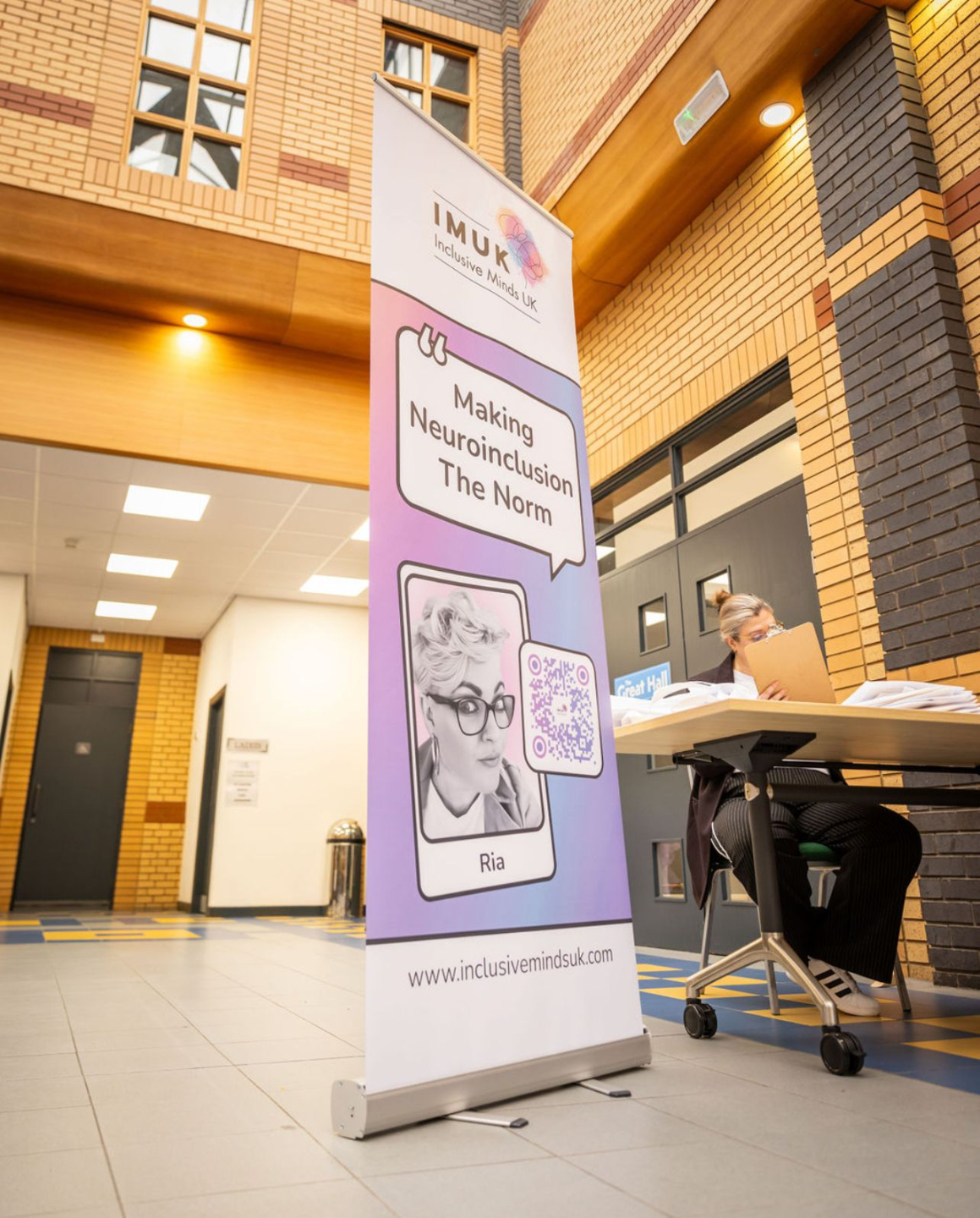 A vertical banner for Inclusive Minds UK (IMUK) stands in a bright indoor space with brick and glass architecture. The banner features a black-and-white photo of Ria, a QR code, and the slogan "Making Neuroinclusion The Norm." A person with short hair and glasses sits at a nearby table, reviewing papers. The table is covered with documents, and a sign in the background reads "Great Hall."