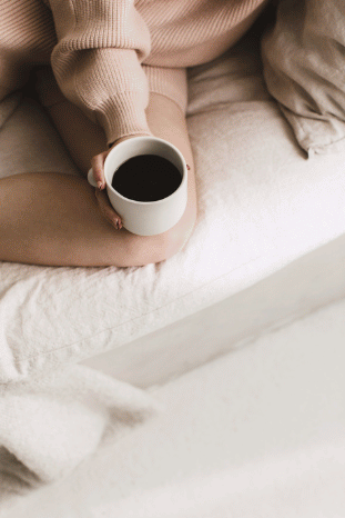 A cozy, aerial view of someone wrapped in a soft blush sweater, safely holding a warm white mug of coffee on clean, comfortable linen sheets