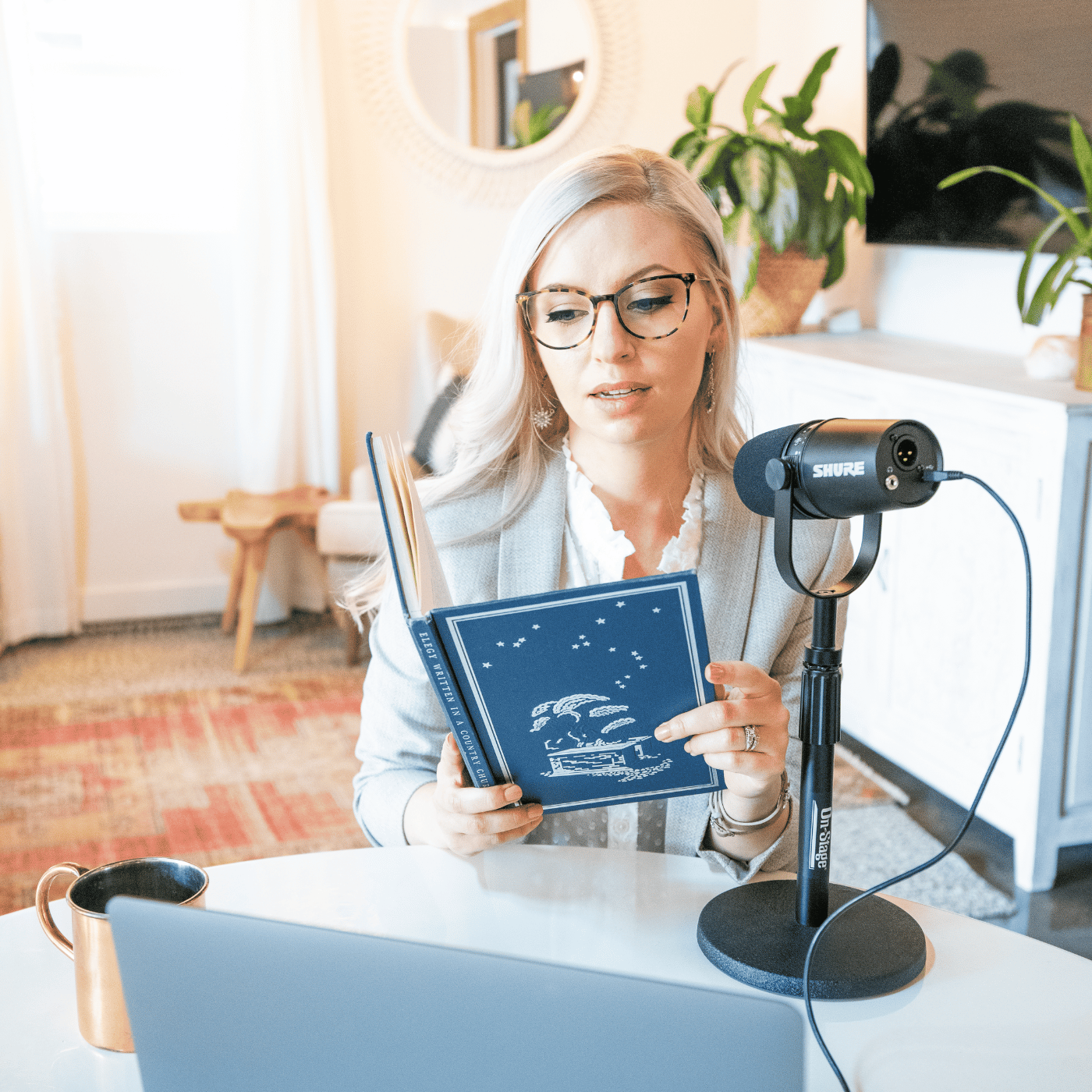 A photo of Jordan Blair. She is wearing a white shirt and light grey blazer. She reads from a blue book into a podcast microphone