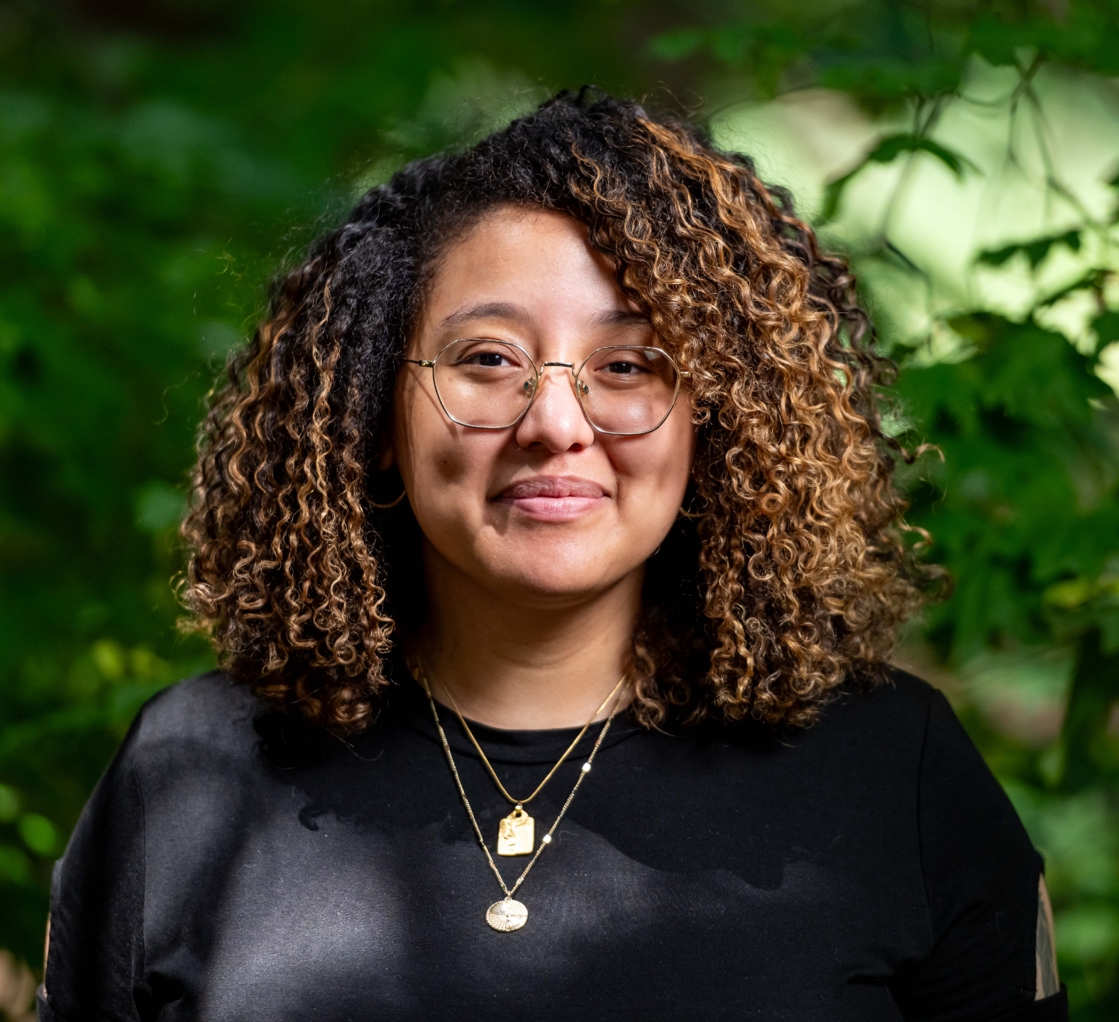Headshot. Ariana Brown, a light skinned Black woman, stands against a tree in a field. She is smiling a closed smile and wears long havana twists and a plaid button down.