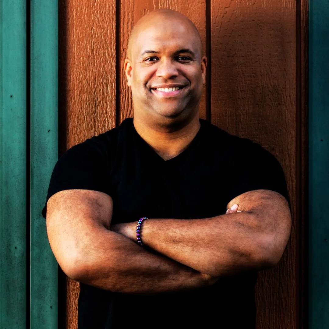 Mike Veny is standing confidently with his arms crossed in front of a wooden wall painted in brown and green. He is wearing a black T-shirt and a beaded bracelet, smiling warmly at the camera. The setting has a natural and relaxed vibe, emphasizing his approachable demeanor.
