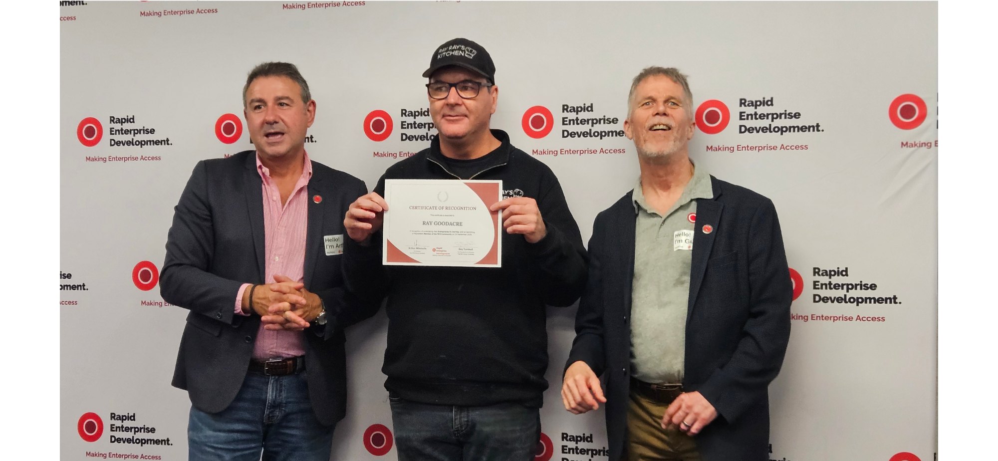 “Ray Ray’s Kitchen’ Founder, Ray Goodacre, a mid-aged white man holds his ‘RED Certificate of Achievement’. He stands in between Dr. Guy Turnbull and Arthur Mitsioulis, the co-founders of The RED Group in Australia. They are mid-aged white males, pictured in front of a wall showing the RED logo at an event for entrepreneurs with lived experience of disability, and those supporting them