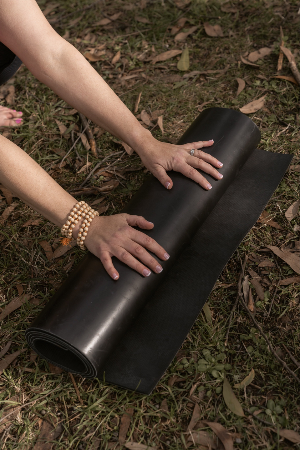 hands one with meditation beadsmwith part of the arms visible resting on a black yoga mat that is resting on grass and leaves