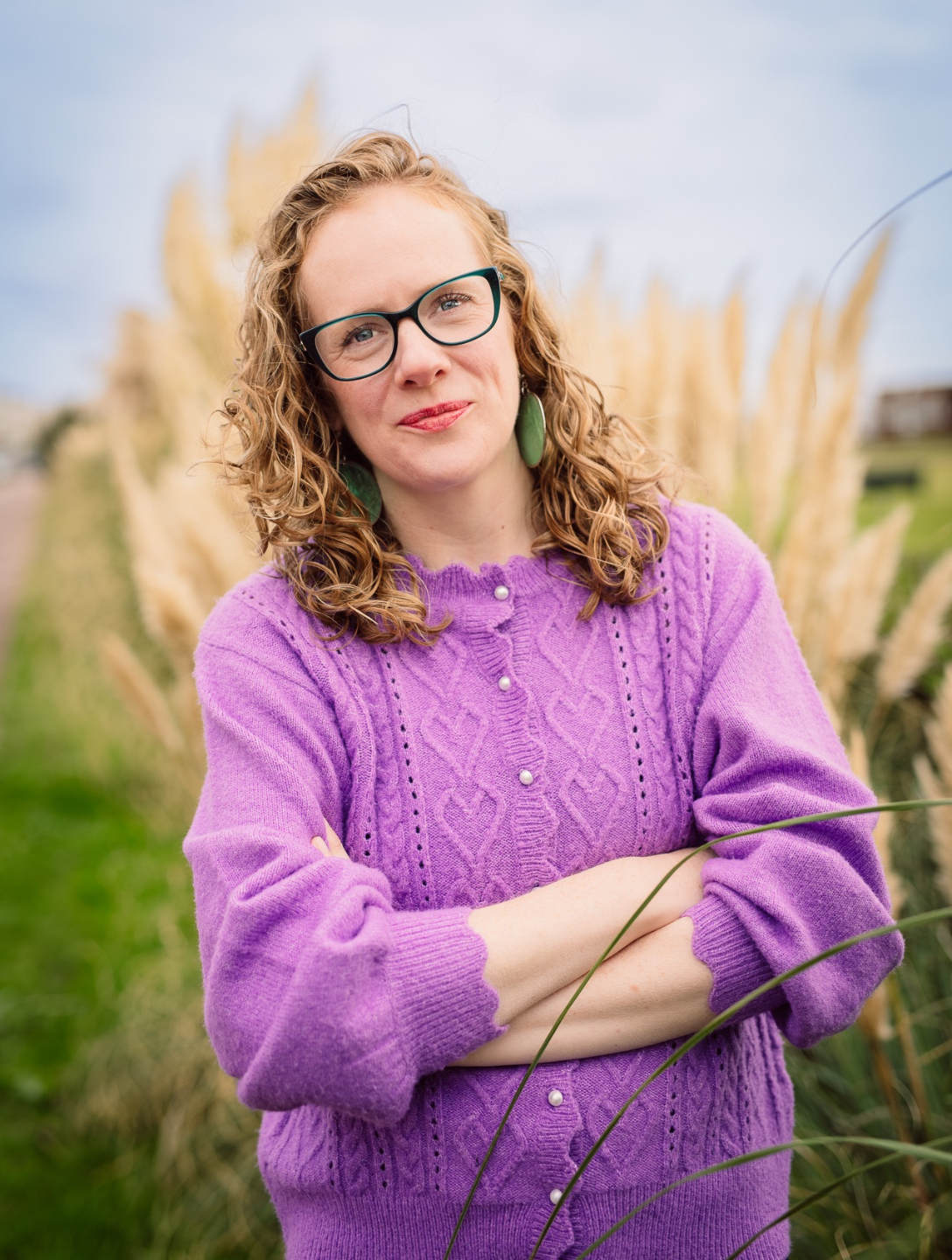 Photo of Jess Meredith wearing a purple cardigan and green framed glasses. She is folding her arms in front of her and her hair is curly and down.
