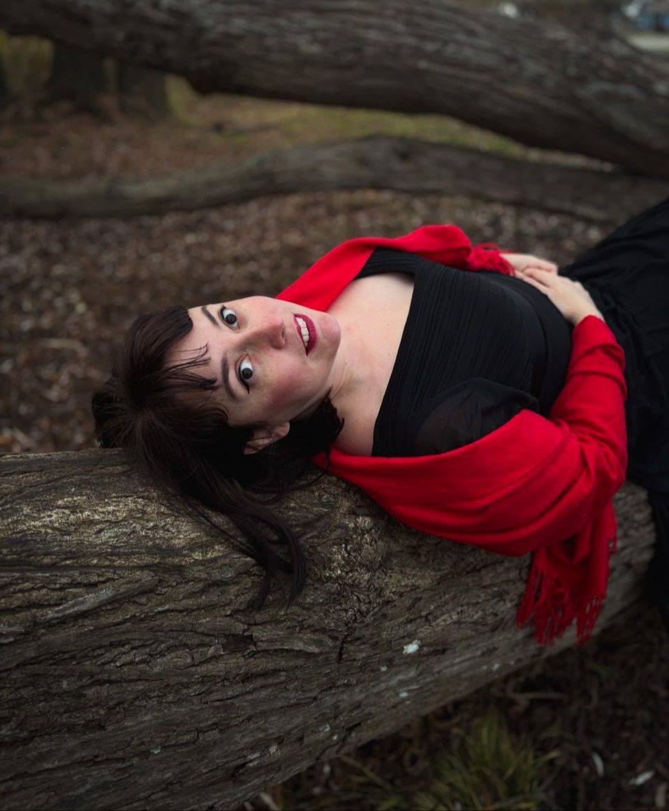 A woman with dark hair wearing a black skirt and white shirt, standing in nature surrounded by autumn trees with orange-red leaves. She appears happy and peaceful.