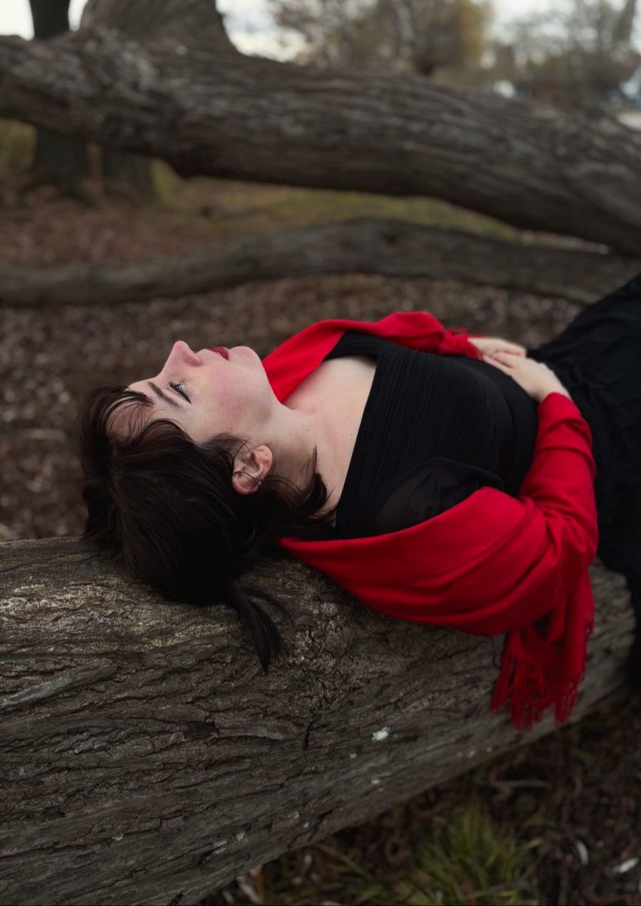 A woman with dark hair wearing a black skirt and white shirt, standing in nature with her hands on her shoulders, surrounded by autumn trees with orange-red leaves.