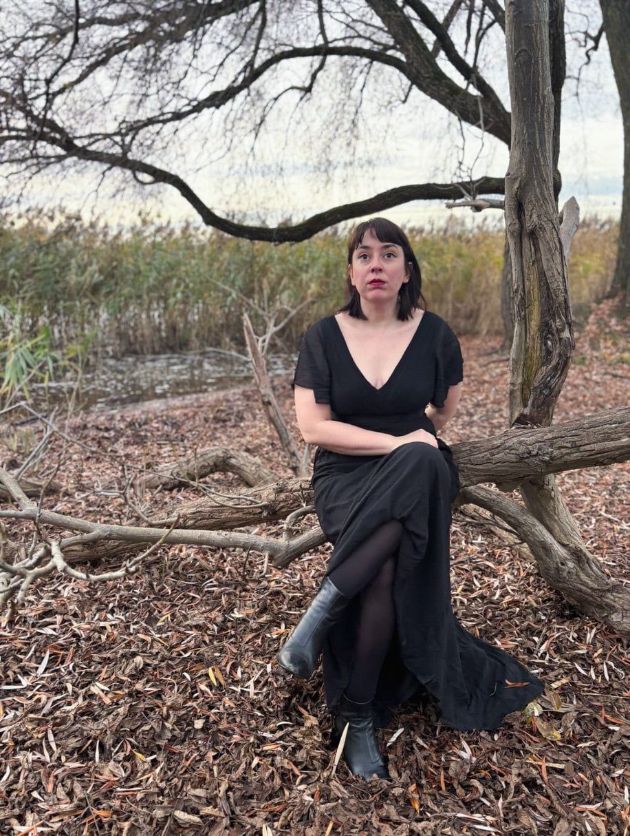 Sophie, founder of Embodied Sacred Feminine Wisdom, sitting between green plants, wearing a blue top and red lipstick, looking at the camera.