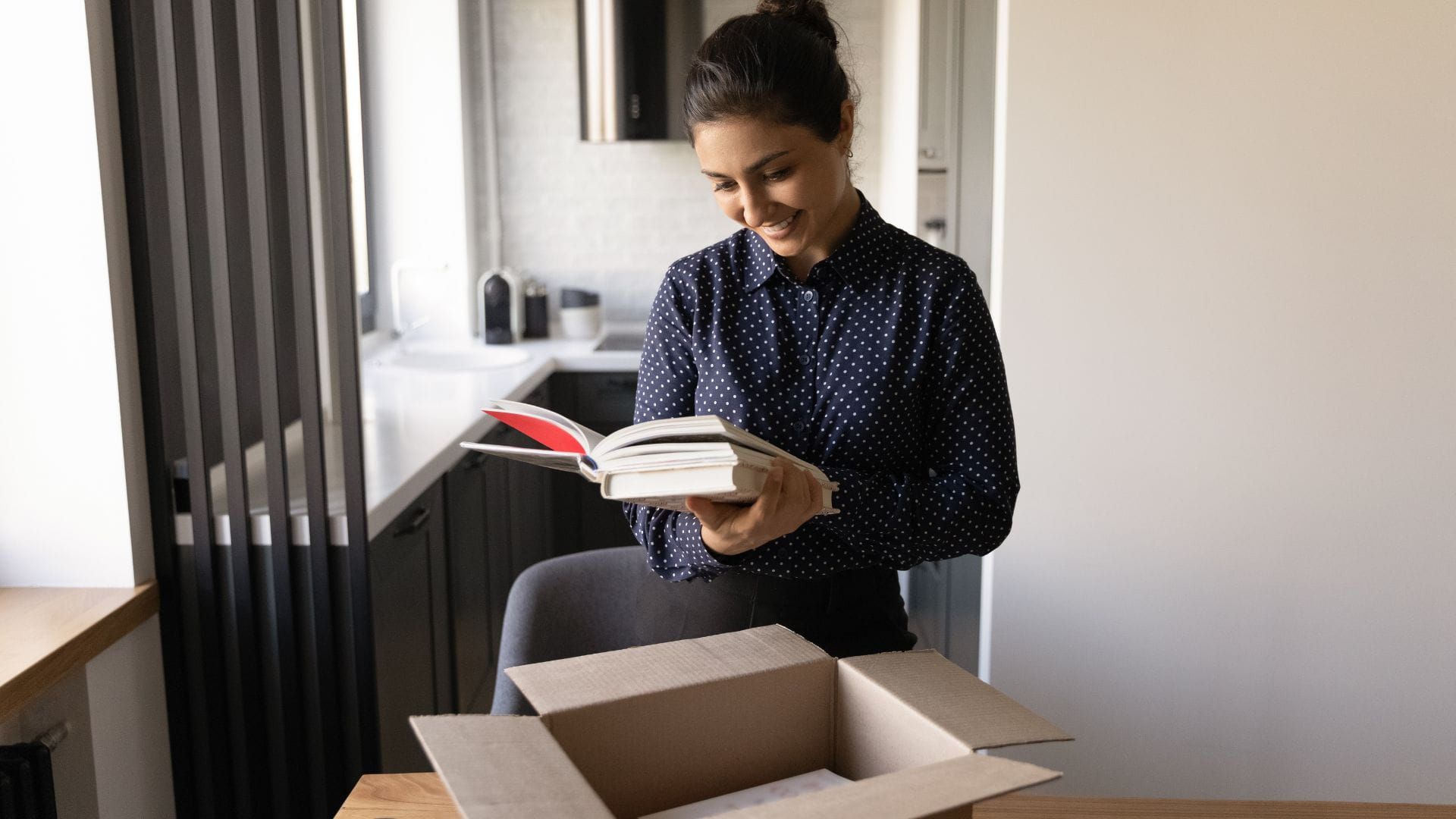 A dark haired woman in a Navy blue and white spotted blouse looks joyfully at her newly authored books that she's taken from a cardboard box on a desk_how to get self published when you're a well-being expert