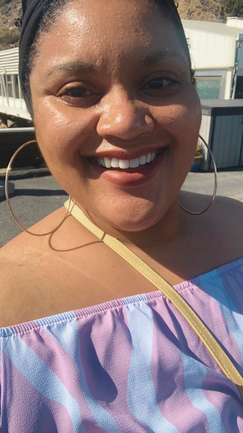 [Image ID] Melinda, a light skinned Black person, is wearing a pink and white off the shoulder striped shirt, a pair of large gold hoop earrings and a nude lipstick. They are smiling warmly and are in the background is a building along the Pacific Coast in Malibu on a sunny day with the mountains in the background