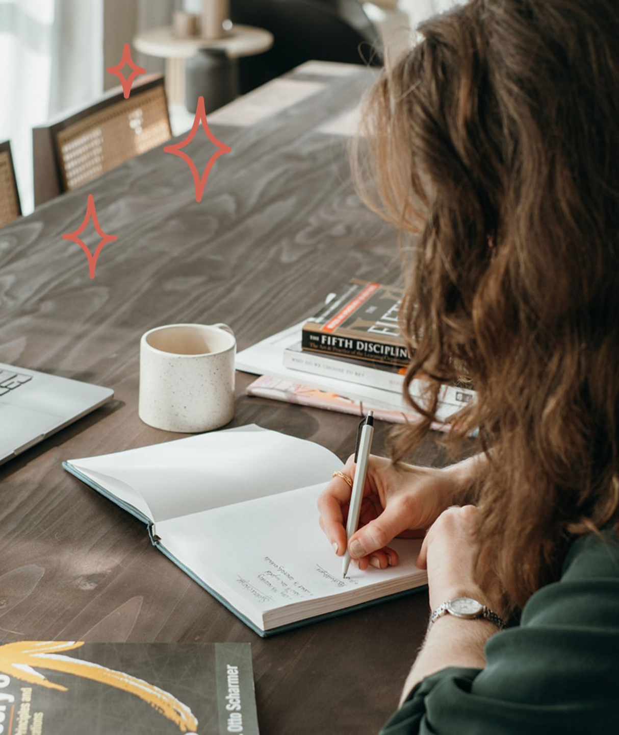 Someone wirting in a notebook while also look at another book. A laptop and coffee mug are on their table.
