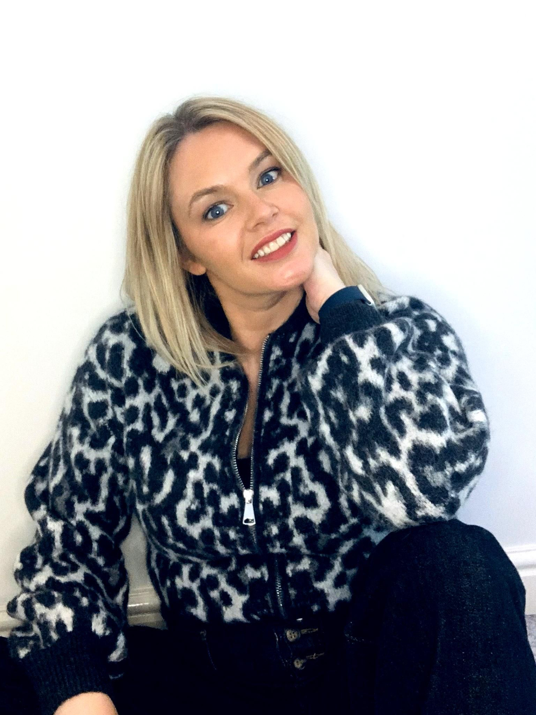 Photo of Emma Smith sitting against a white wall, smiling with her head tilted and wearing a grey and black leopard-print zip-up jumper.