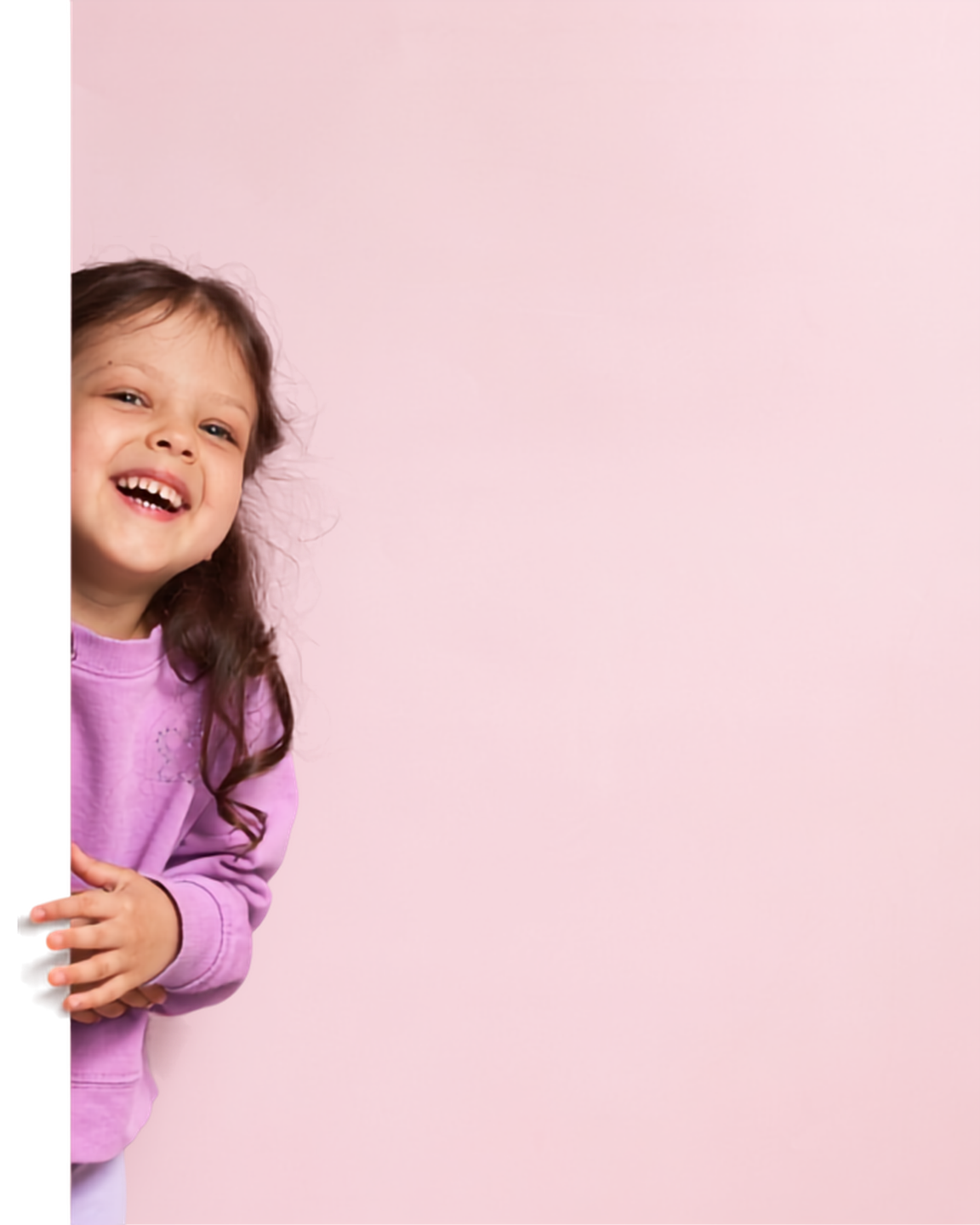 A preschool-aged girl in a purple jumper leaning around a wall, laughing
