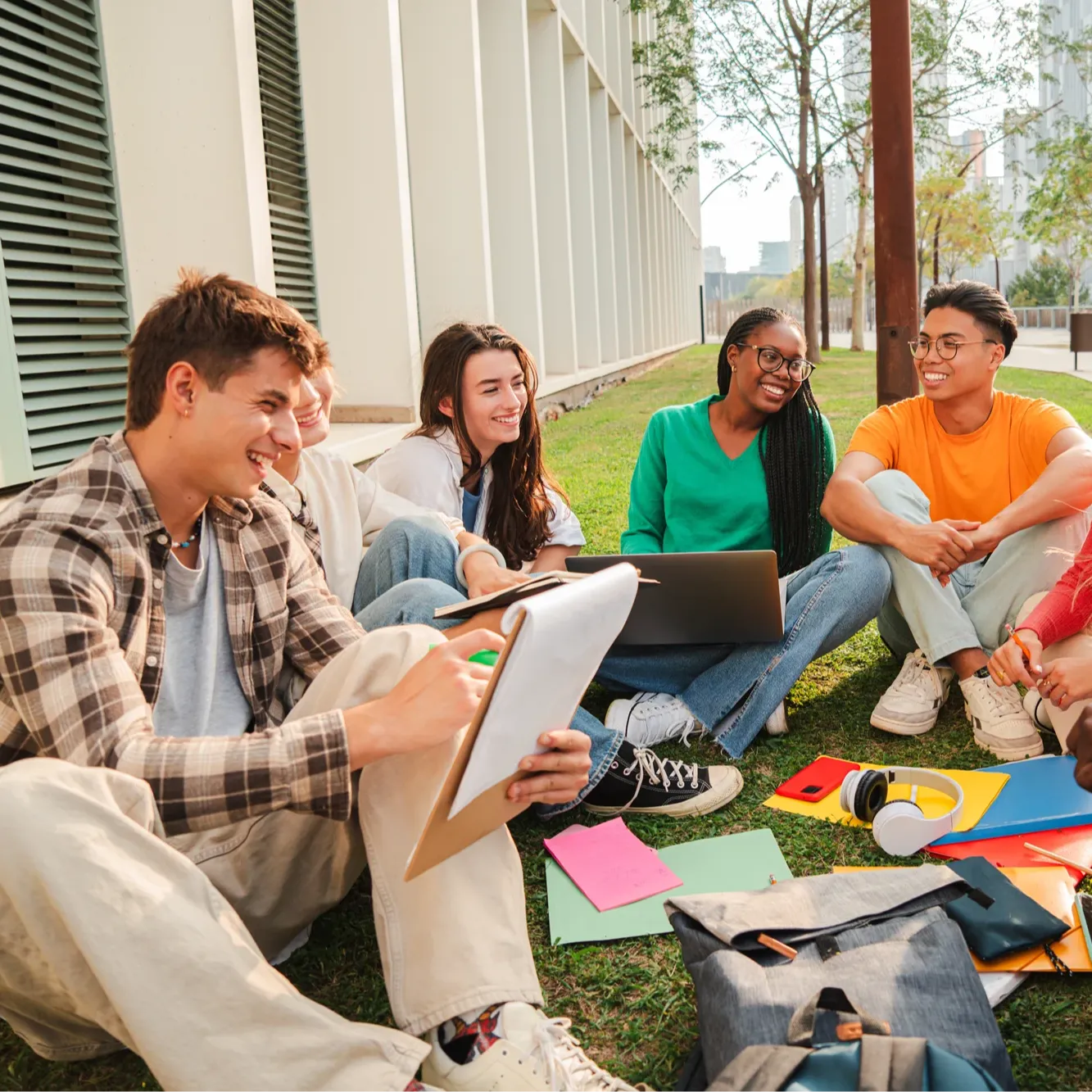 Five young adults sit on grass outside a building, smiling and studying together with notebooks and a laptop. They appear engaged and happy, supporting each other’s mental health during a relaxing group study session on a sunny day.