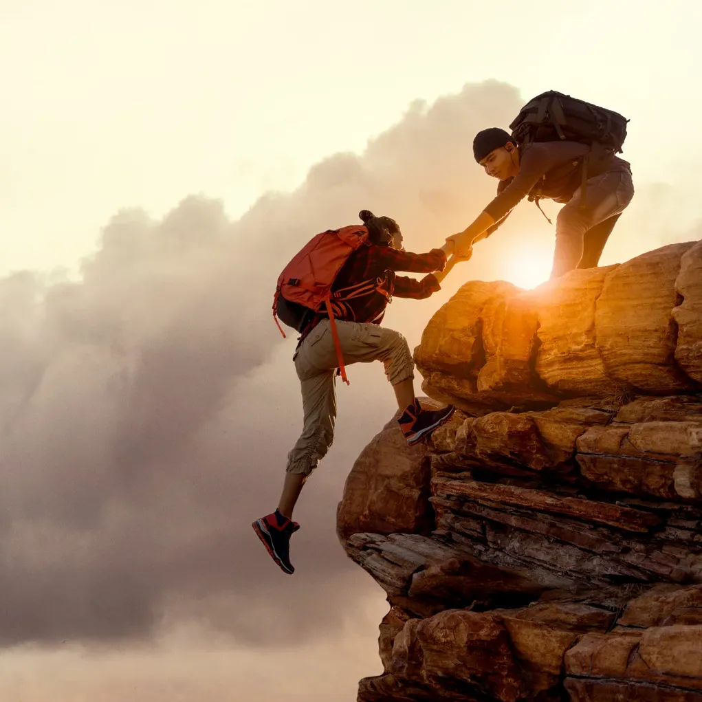 A person with a backpack helps another climb up a rocky cliff at sunset, dramatic clouds and sunlight shining between them—a powerful reminder of the importance of support in mental health journeys.