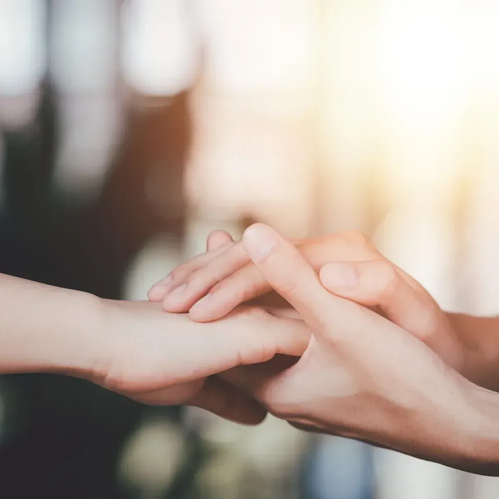 Two hands gently holding another person's hand, conveying comfort, support, or compassion—symbolizing First Aid for Mental Health—bathed in a soft, warm light in the background.