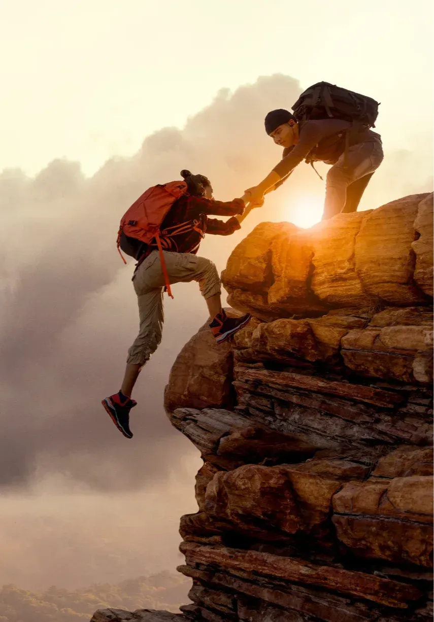 A person offers First Aid for Mental Health by supporting another to climb a rocky cliff at sunset, both in outdoor gear and backpacks, sunlight shining between them against a backdrop of clouds.