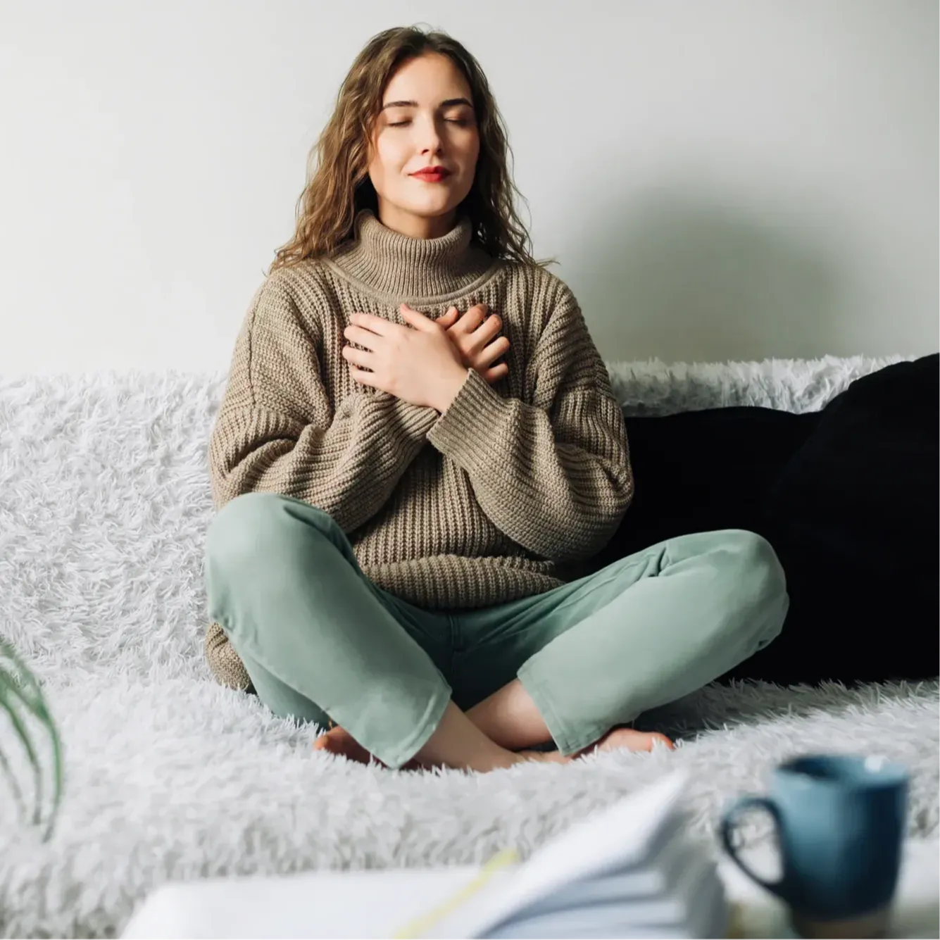 A woman with long hair sits cross-legged on a couch, eyes closed and hands resting on her chest. She wears a brown sweater and light green pants, practicing First Aid for Mental Health in a calm, cozy, and peaceful setting.
