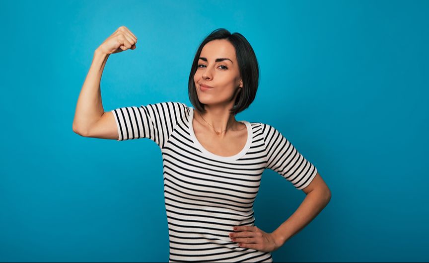 A woman with dark hair and a white shirt with black stripes has one arm on her hip and the other up in a fist showing her strong arm muscles, with a confident look on her face.