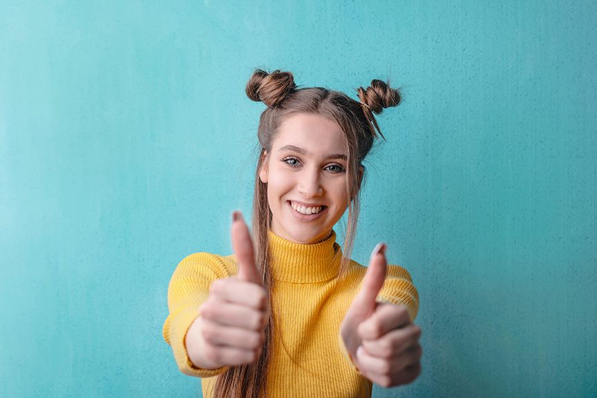 A woman with blonde hair and a yellow turtleneck sweater is holding up two thumbs and smiling.