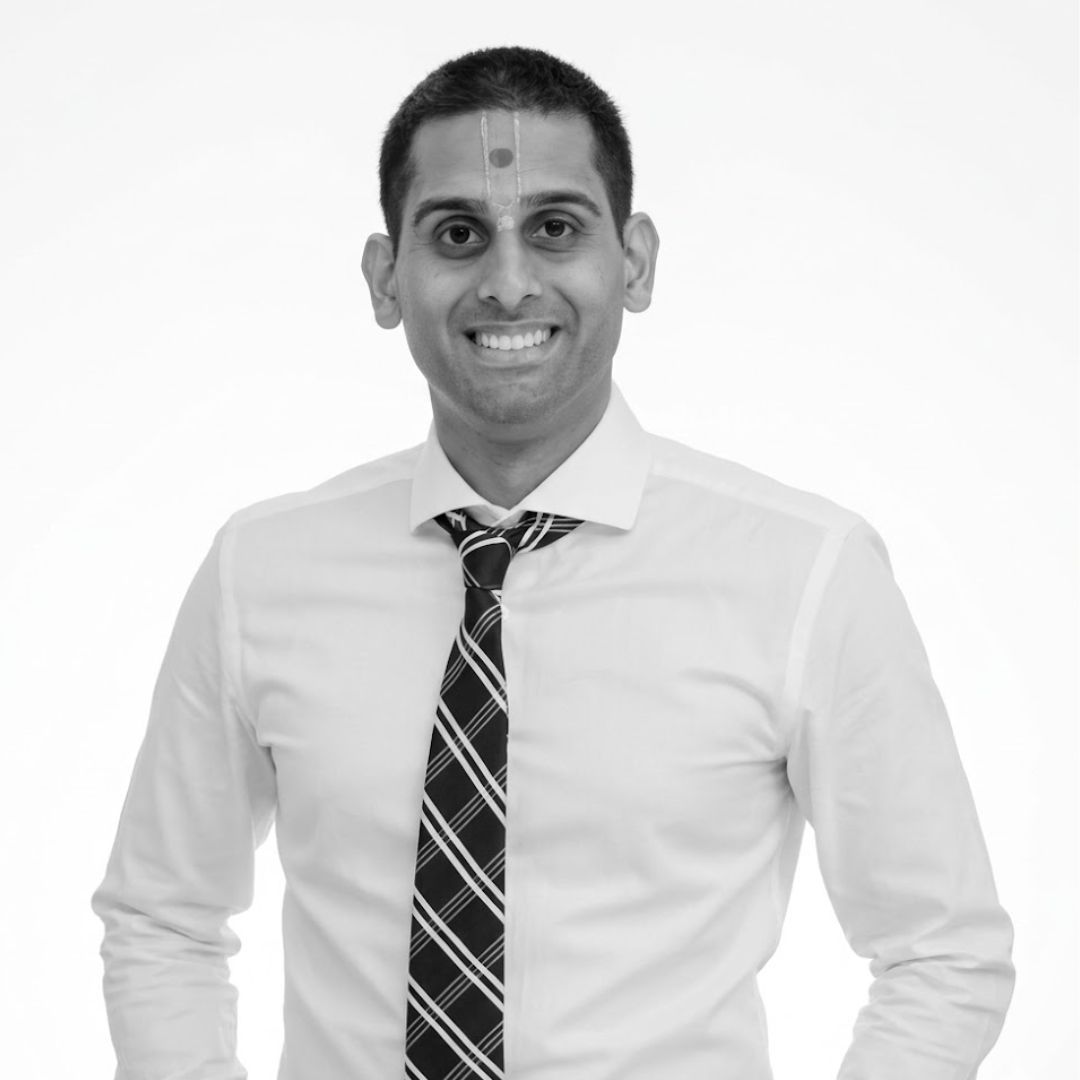 Smiling man with short hair wearing a white dress shirt and patterned tie, with a forehead marking, facing forward against a plain background