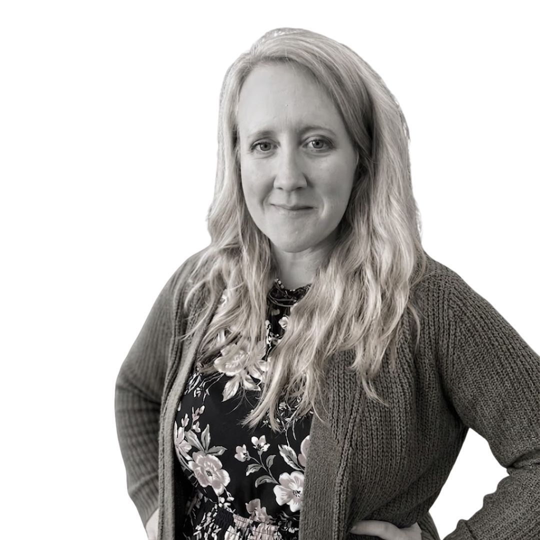 Smiling woman with long wavy hair wearing a floral top and knit cardigan, standing with hands on hips against a plain background