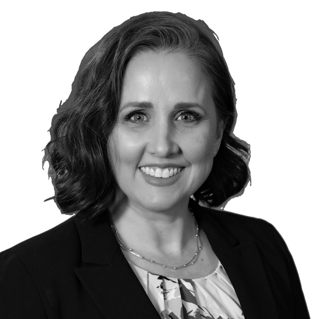 Smiling woman with shoulder-length wavy hair wearing a blazer, patterned blouse, and necklace, facing forward against a plain background