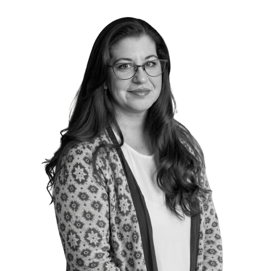 Smiling woman with long wavy hair wearing glasses and a patterned cardigan over a light top, facing forward against a plain background