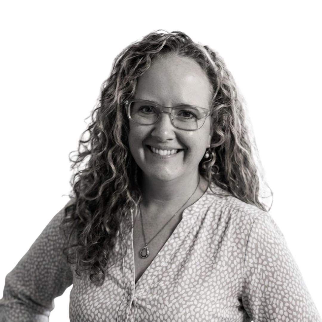 Smiling woman with curly hair wearing glasses and a patterned blouse, facing forward against a plain background