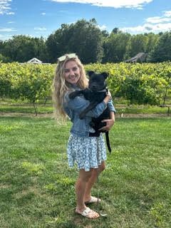 Handler is wearing a blue floral dress while holding a medium sized shorthair black pup. They are both looking into the camera smiling.