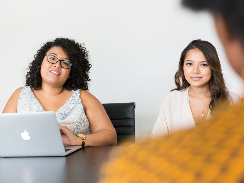 Two women in a relaxed business meeting, smiling and listening attentively to the person opposite, representing open communication, confidence, and connection.