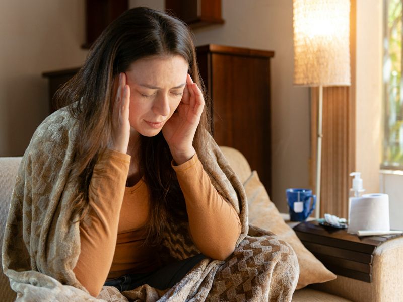 Woman sitting on a sofa holding her head in discomfort, representing headache and migraine relief through calm relaxation, stress reduction, and hypnotherapy - Retrained Minds Hypnotherapy.