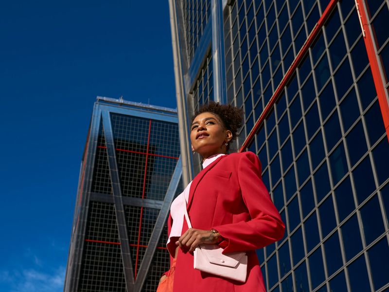Confident woman in a red suit standing tall before modern glass buildings, symbolising self-belief, ambition, and professional growth - Retrained Minds Hypnotherapy.