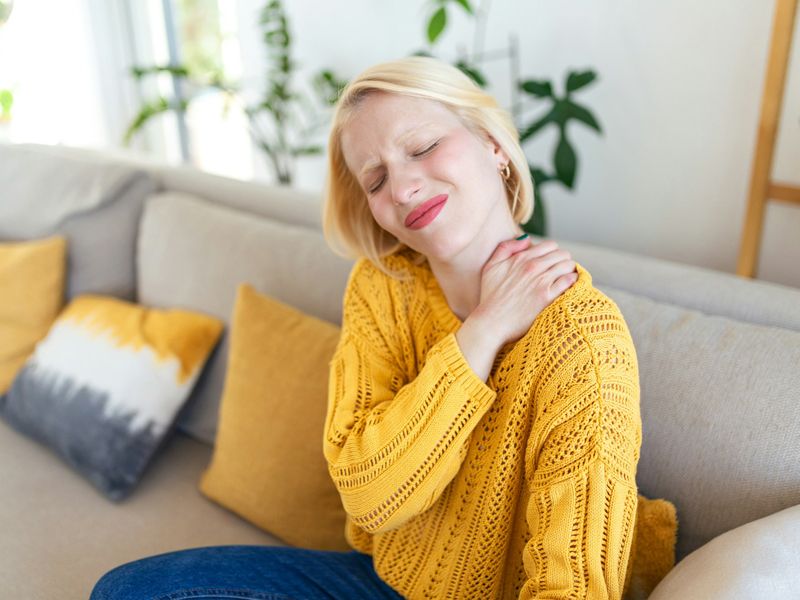 Woman sitting on a sofa gently holding her neck and shoulder in discomfort, representing relief from chronic pain, stress release, and natural healing through hypnotherapy - Retrained Minds Hypnotherapy.