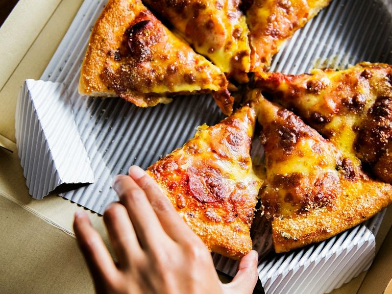 Hand reaching for a slice of pizza, symbolising emotional eating, cravings, and the challenge of maintaining control - Retrained Minds Hypnotherapy.
