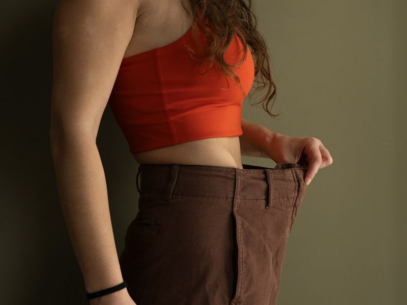 Person in a red top showing weight loss progress by holding out loose trousers, symbolising achievement, confidence, and healthy transformation - Retrained Minds Hypnotherapy.